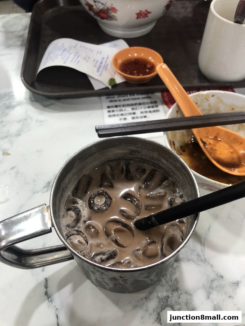 A close-up of a metal mug filled with a cold beverage and ice, with chopsticks and a small dish of sauce.