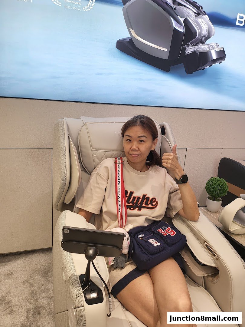 A woman sits in a massage chair, giving a thumbs-up, with a large screen displaying a massage chair behind her.