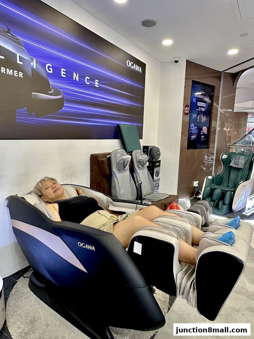 A person relaxing in an OGAWA massage chair with other chairs and a promotional display in the background.