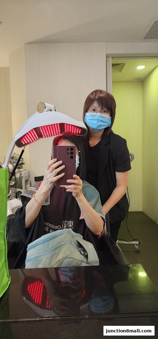 A client receives a hair treatment under a red light therapy device, with a stylist standing behind.