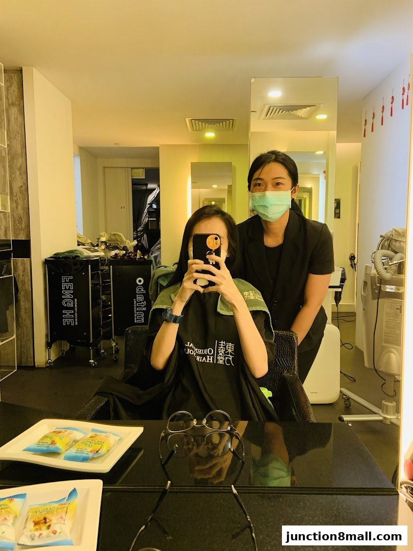 A woman in a salon chair takes a selfie with a stylist standing behind her.