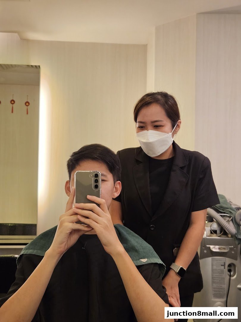 A hairstylist in a black jacket and mask stands behind a client taking a selfie.