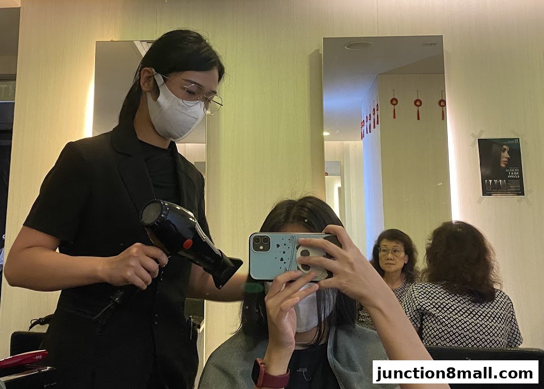 A hairstylist dries a client's hair while the client takes a selfie in the mirror.