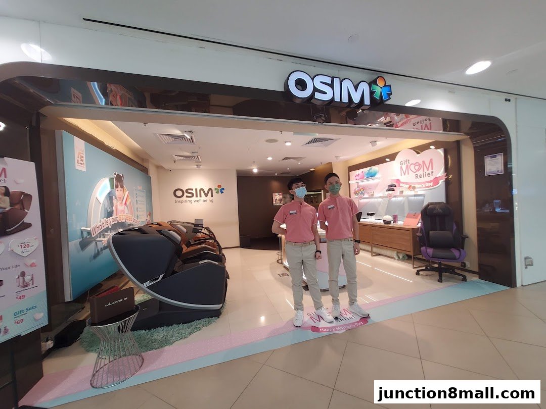 Two staff members stand in front of the OSIM shop, showcasing massage chairs and wellness products.