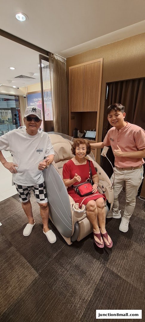 A smiling couple tries out a massage chair with a staff member giving a thumbs up.