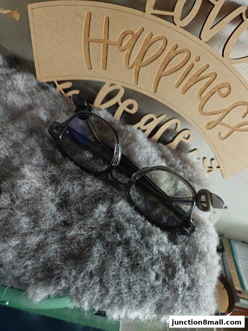 A pair of stylish eyeglasses rests on a soft, grey textured surface with a decorative wooden sign in the background.