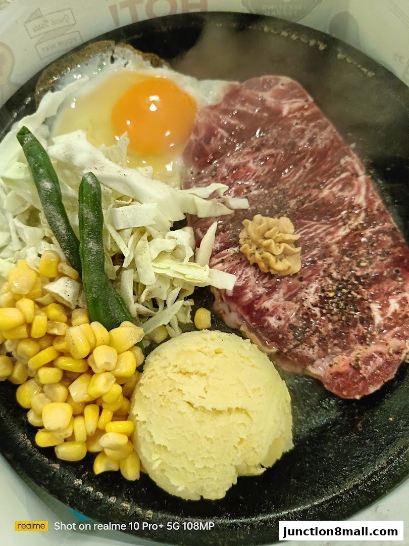 A sizzling steak dish served on a hot plate with a fried egg, corn, mashed potatoes, and vegetables.