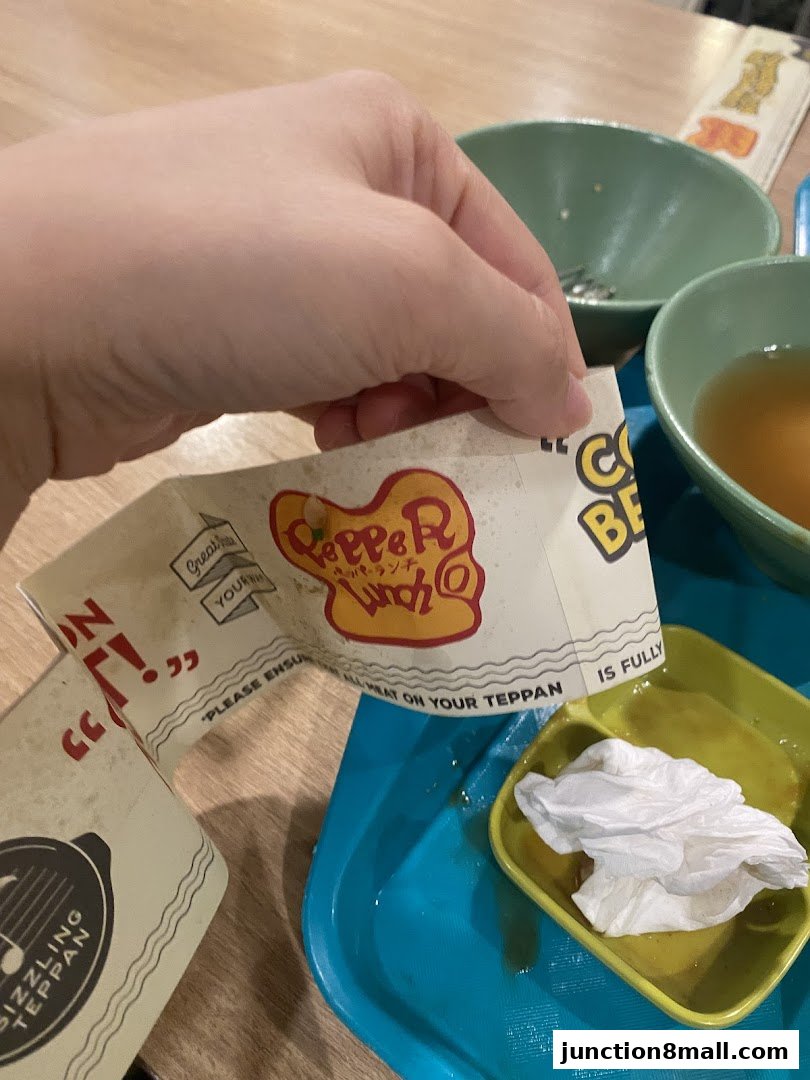 A hand holds a Pepper Lunch Express paper wrapper with food and bowls in the background.