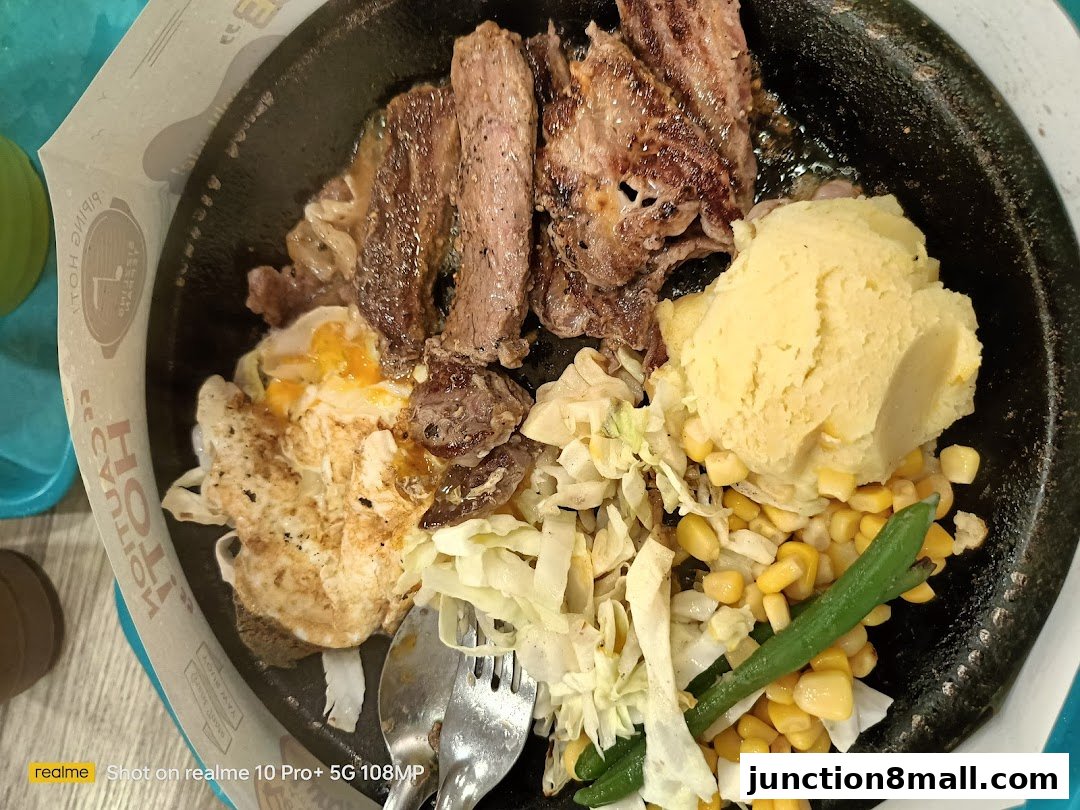 A sizzling steak dish with mashed potatoes, corn, beans, shredded cabbage, and a fried egg.