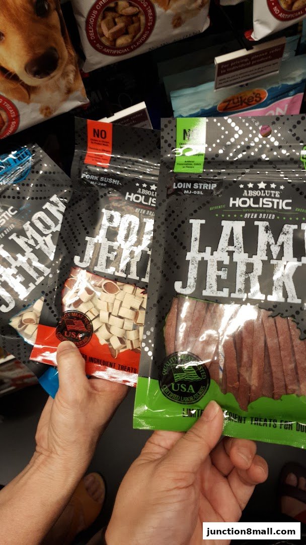Hands holding bags of dog jerky treats, including pork sushi and lamb.