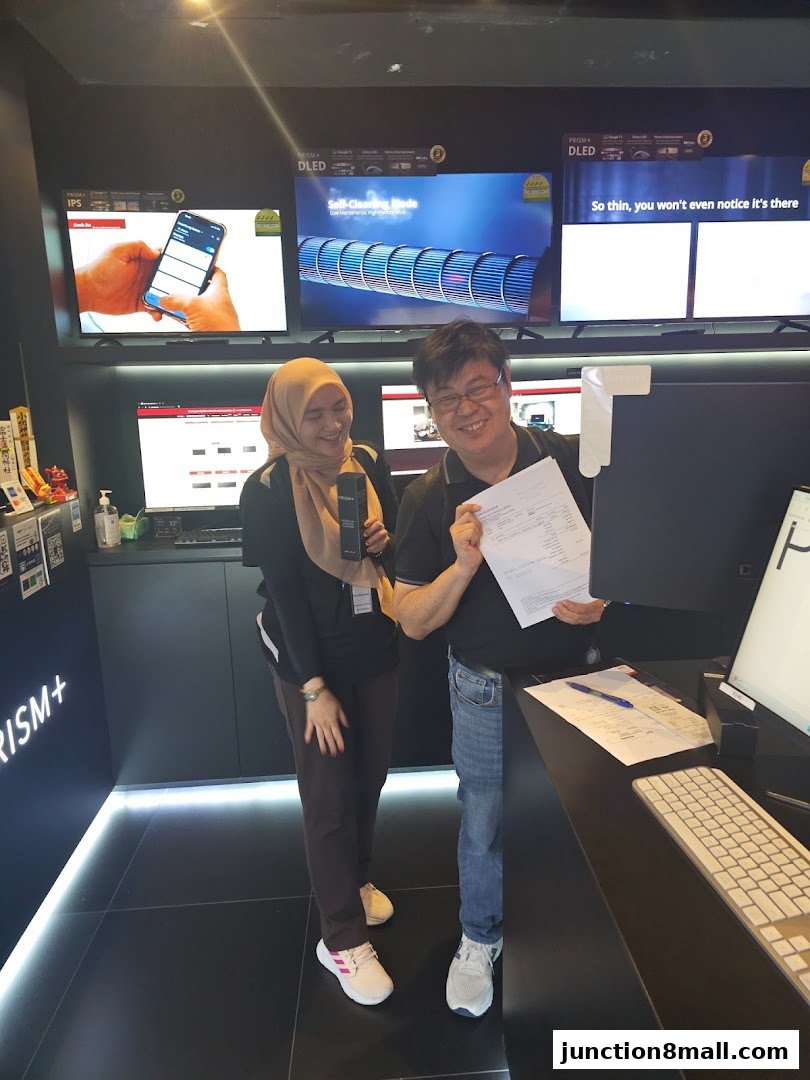 Two smiling people stand in a modern electronics store, with large displays showing TV technology.