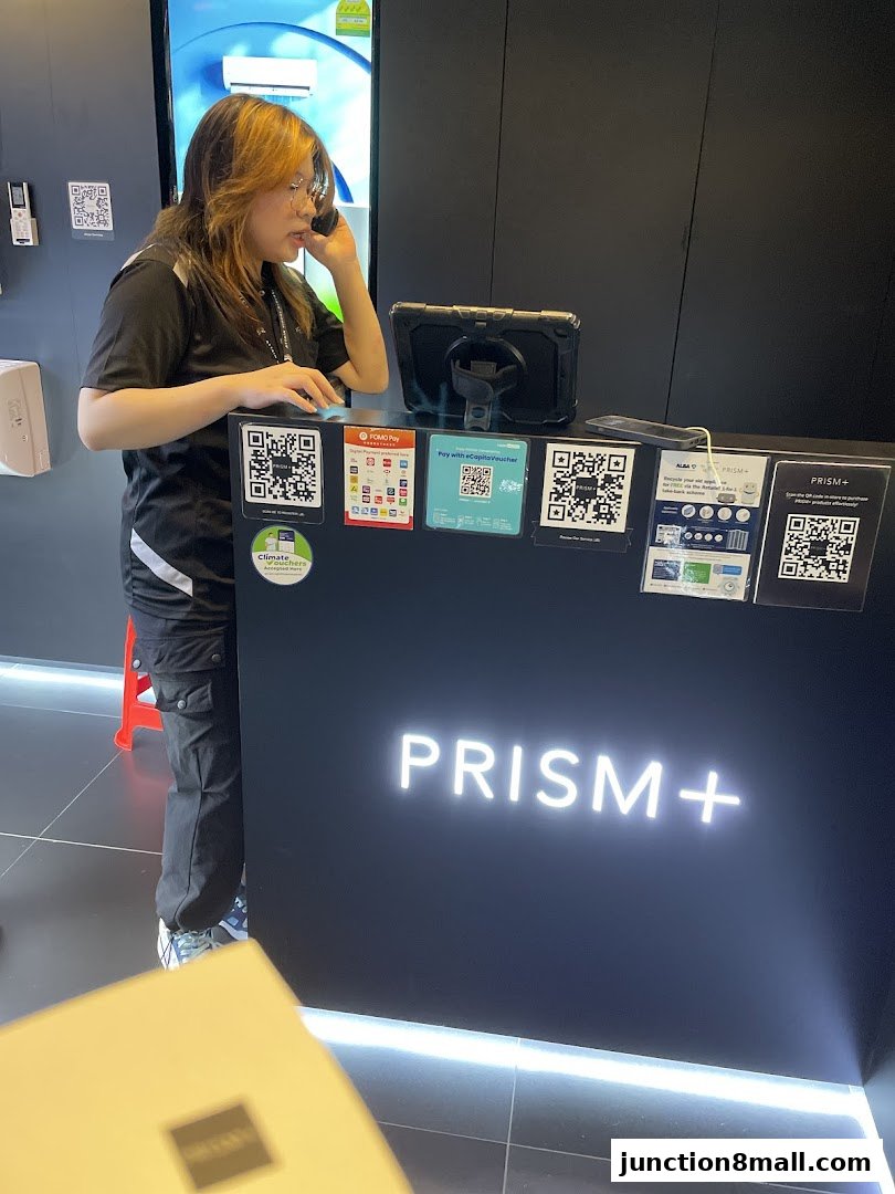 A staff member at the PRISM+ counter, surrounded by payment QR codes and product information.