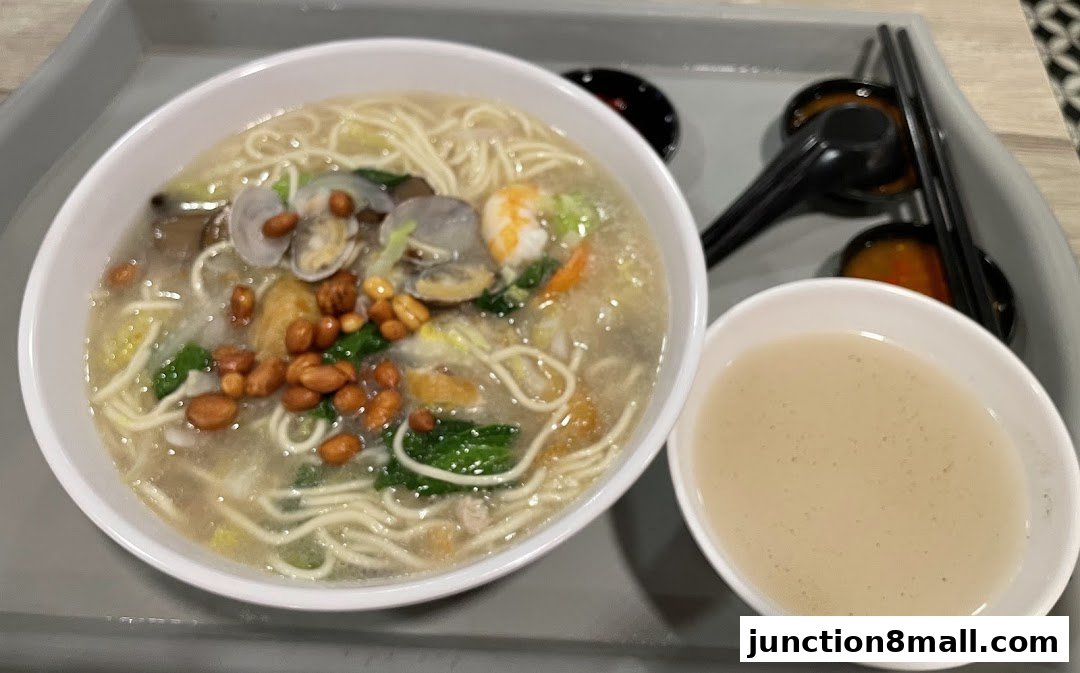A bowl of noodle soup with clams, shrimp, peanuts, and vegetables, served with a side of broth.