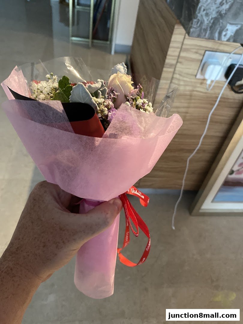 A hand holds a bouquet of flowers wrapped in pink paper and tied with a red ribbon.