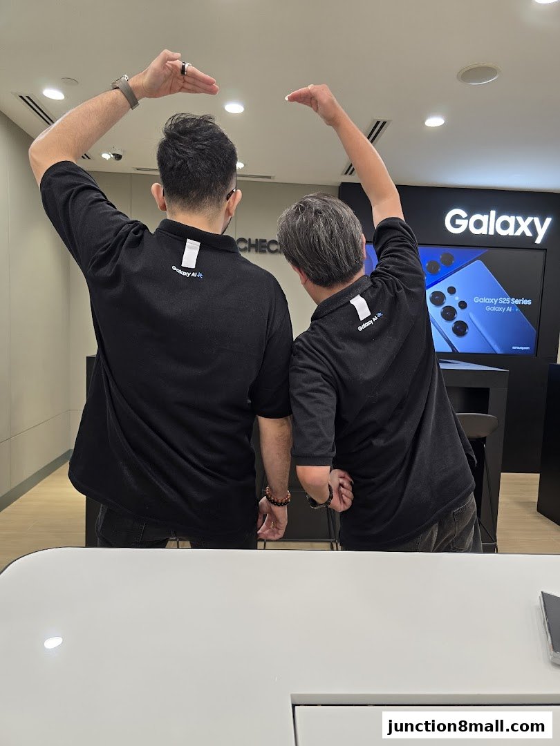 Two staff members in black shirts pose with hands above their heads in a Samsung store.