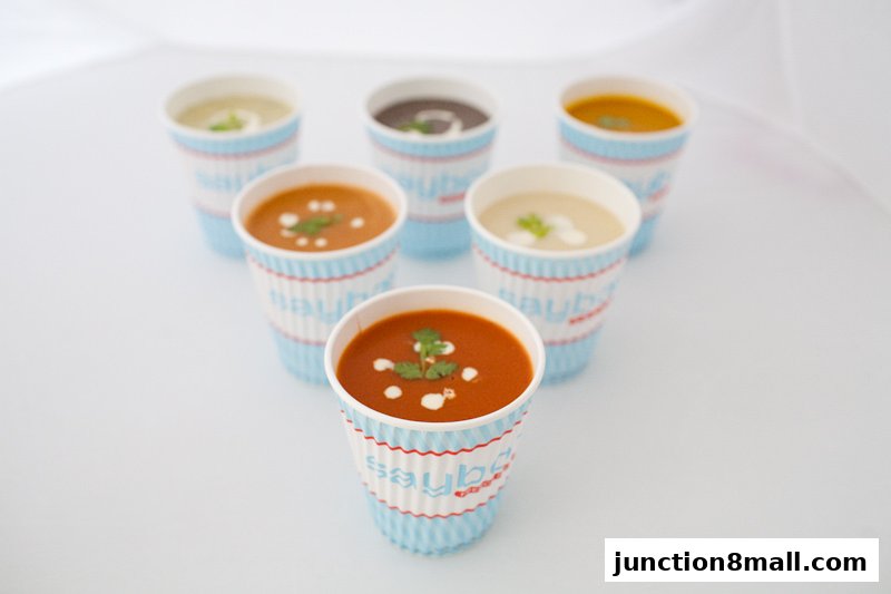 A selection of six colorful soups served in branded cups, garnished with cream and herbs.