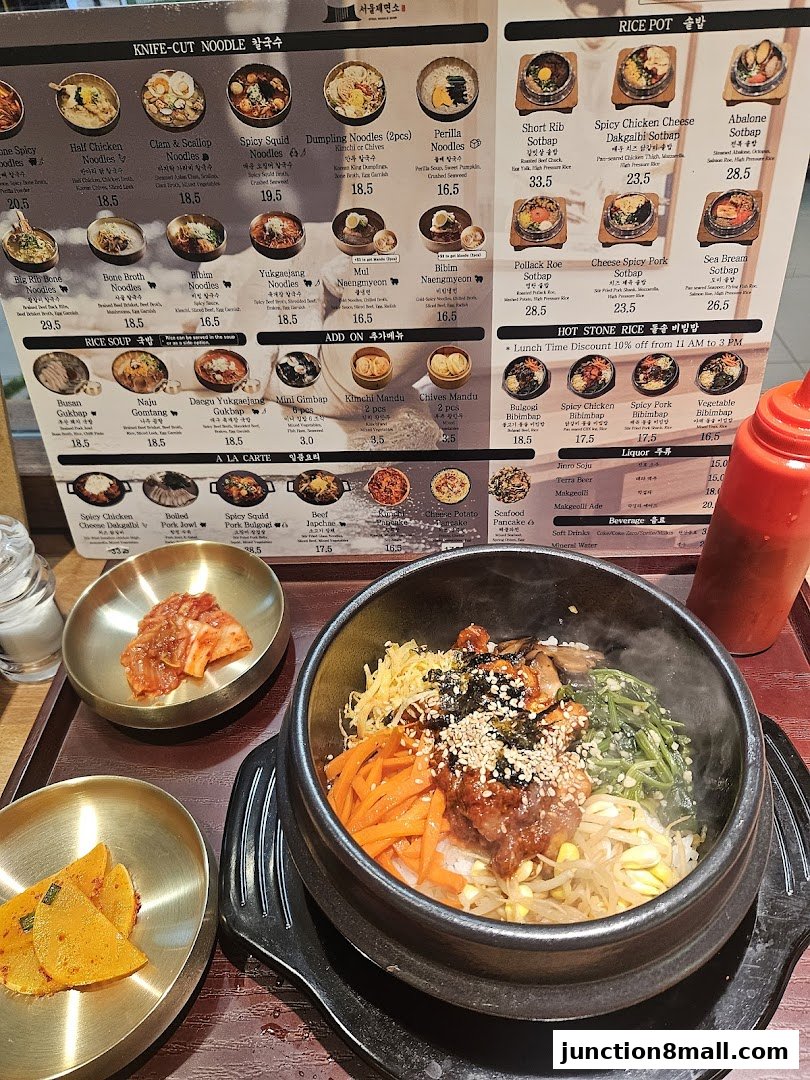 A steaming bowl of bibimbap with various side dishes and a menu displaying Korean noodle and rice dishes.