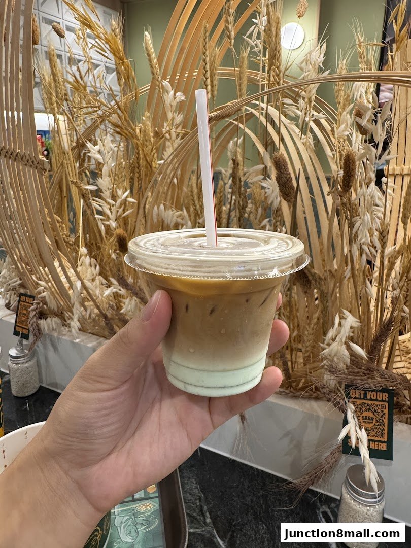 A hand holds a layered iced coffee drink with a straw in front of a decorative wheat display.