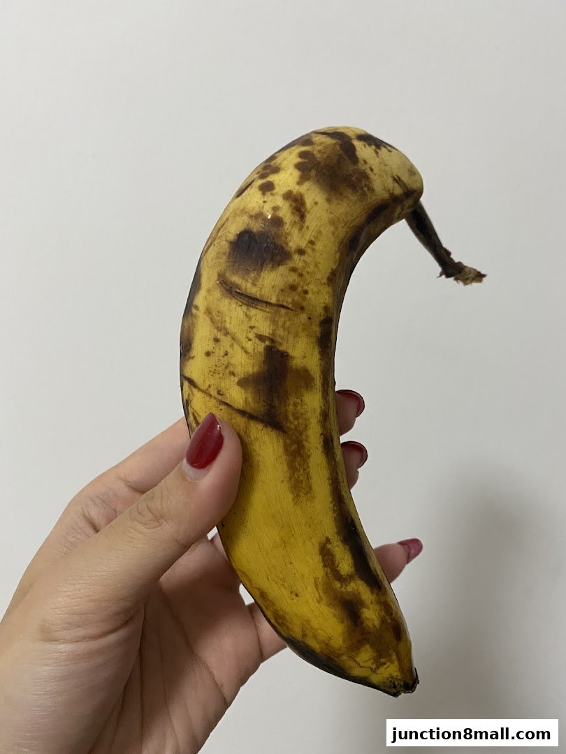 A hand with red nail polish holds a ripe banana with brown spots against a white background.
