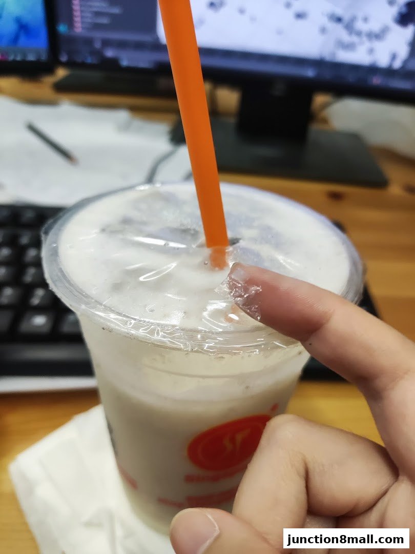 A close-up of a milky beverage with an orange straw and a finger peeling the plastic lid.