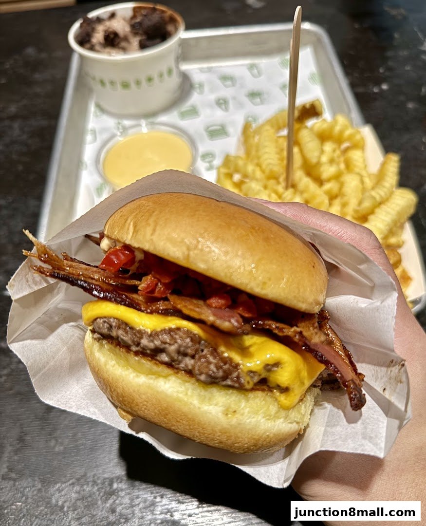 A hand holds a delicious bacon cheeseburger with fries and a shake on a tray.