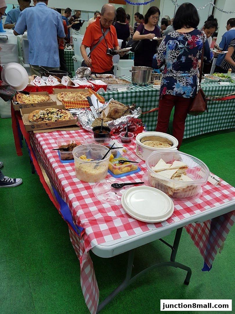 People serving themselves food from a buffet table with pizzas, a roasted turkey, and sandwiches.