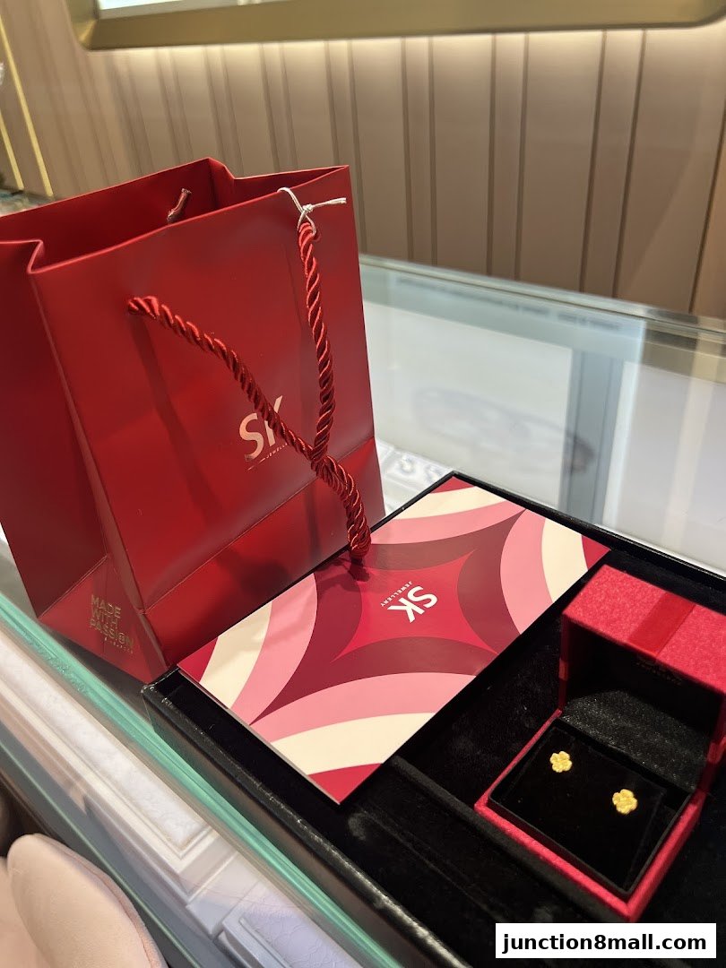 A red SK Jewellery shopping bag with a jewelry box containing gold flower earrings.