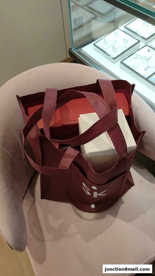 A maroon tote bag with the SK Jewellery logo sits on a pink chair next to a display case.