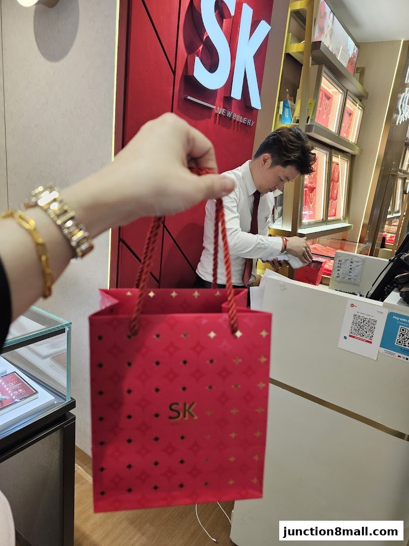 A person holds a red SK Jewellery shopping bag in front of a store counter.