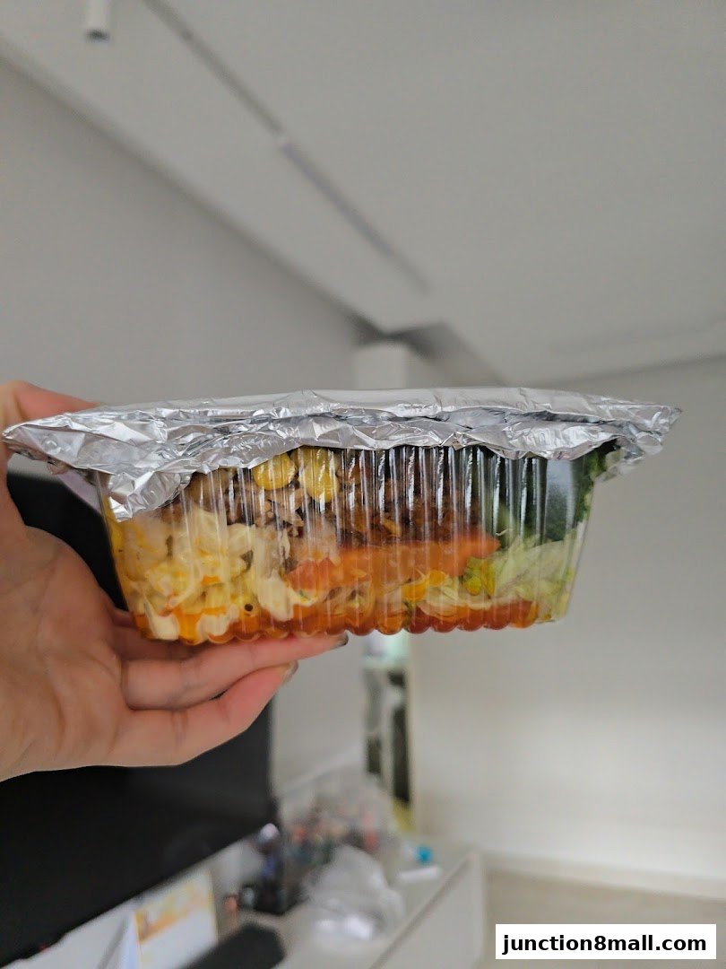 A hand holds a clear plastic container filled with layered food, sealed with foil.