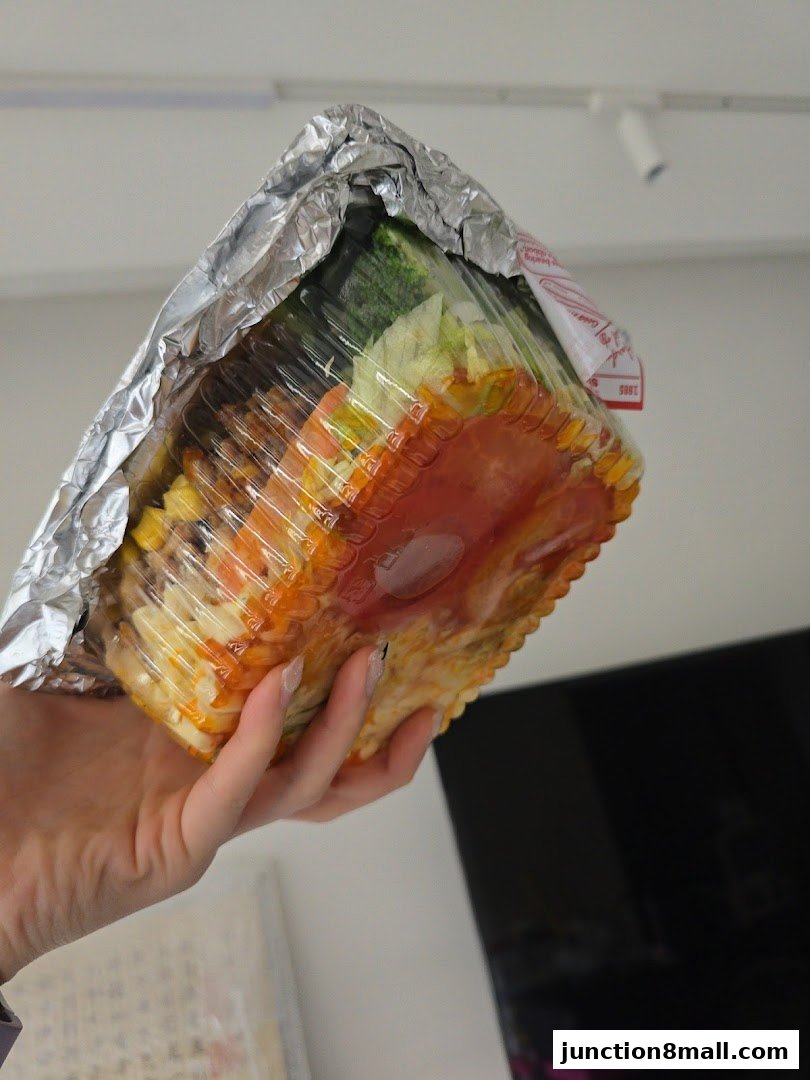 A hand holds a takeout container filled with food, wrapped in foil.