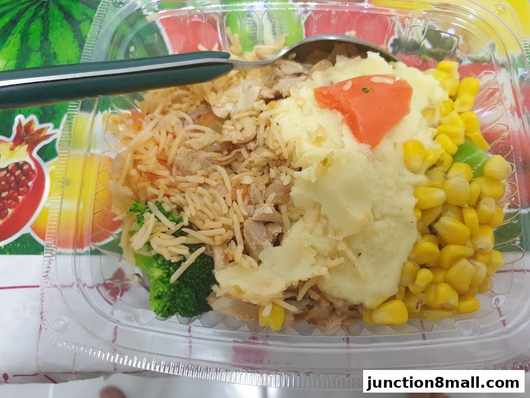 A clear plastic container filled with a meal including rice, mashed potatoes, corn, broccoli, and meat.