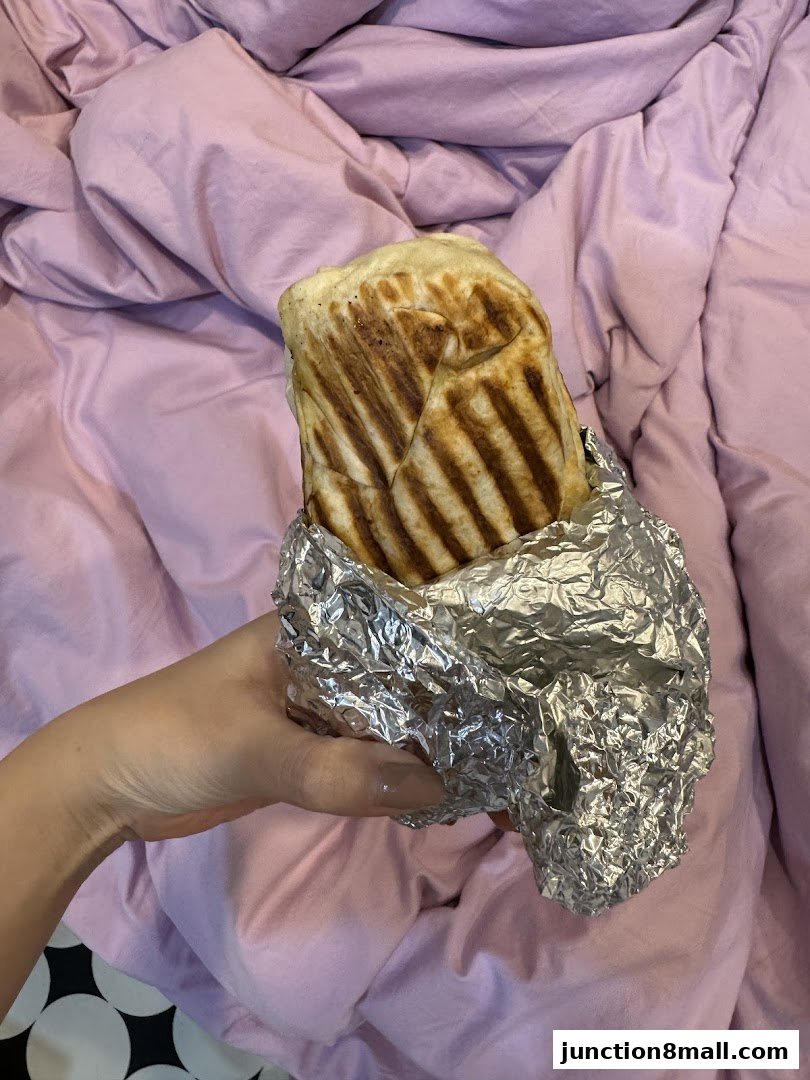 A hand holds a foil-wrapped grilled wrap against a pink, textured background.