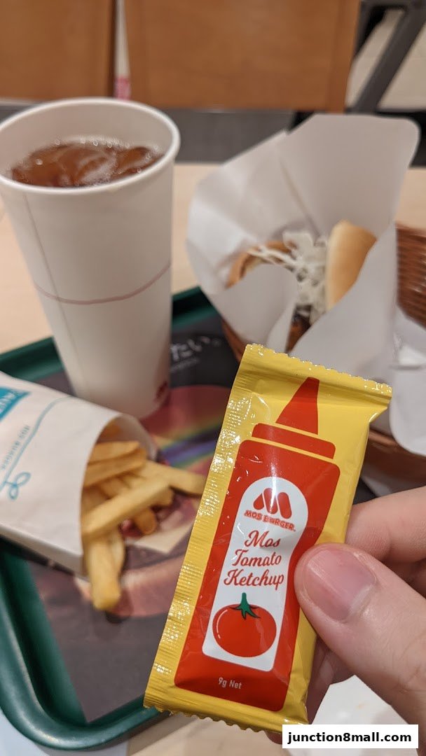 A hand holds a packet of Mos Burger tomato ketchup next to fries, a drink, and a burger.