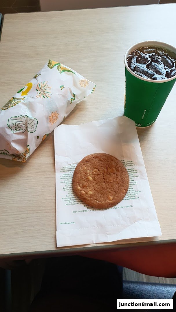 A Subway sandwich, a cookie, and a drink with ice on a wooden table.
