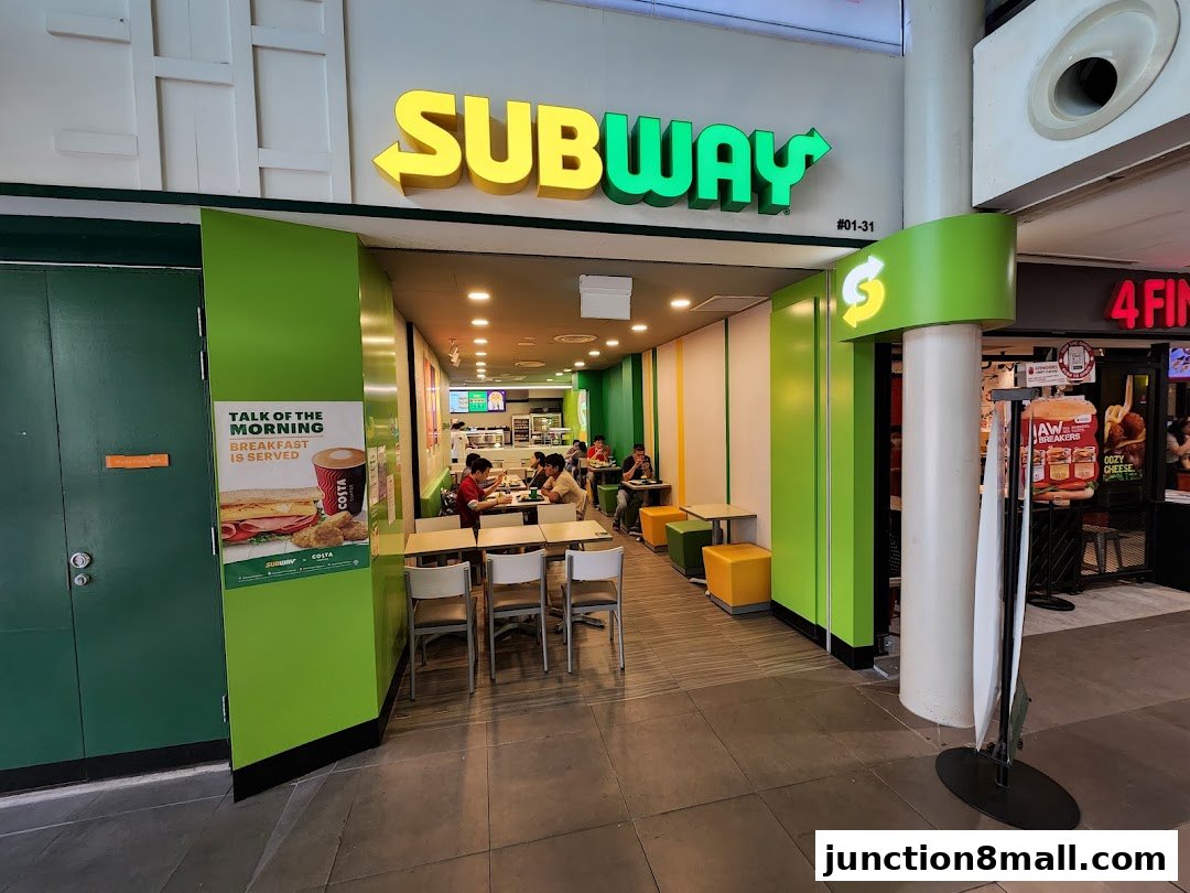 The exterior of a Subway restaurant with its iconic signage and seating area visible.