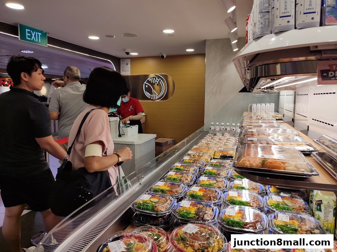 Customers queue at a sushi restaurant with pre-packaged meals displayed in a refrigerated counter.