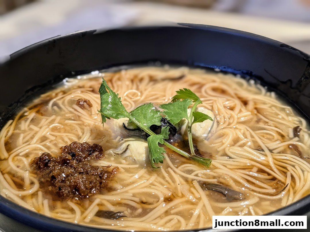 A close-up shot of a steaming bowl of Taiwanese oyster noodles with cilantro and chili paste.