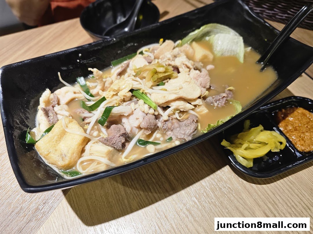 A close-up of a hearty bowl of noodles with meat, tofu, and vegetables, served with side condiments.