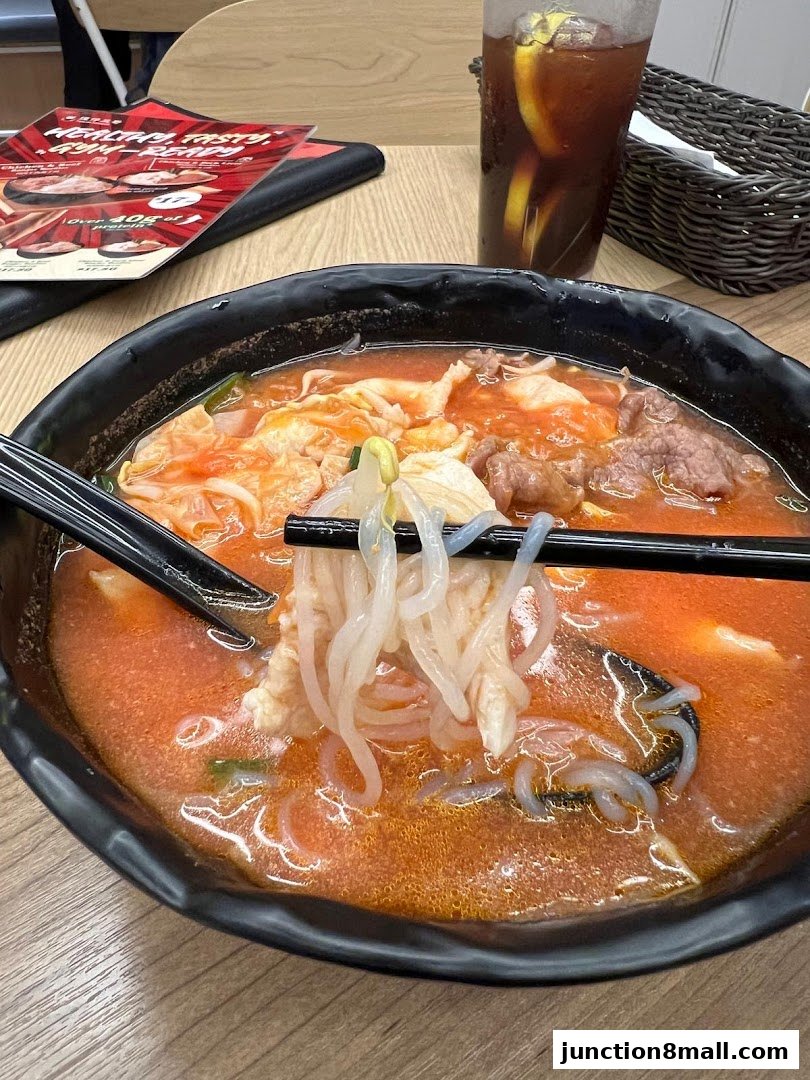 A close-up of a steaming bowl of noodles with meat and vegetables, accompanied by iced tea.