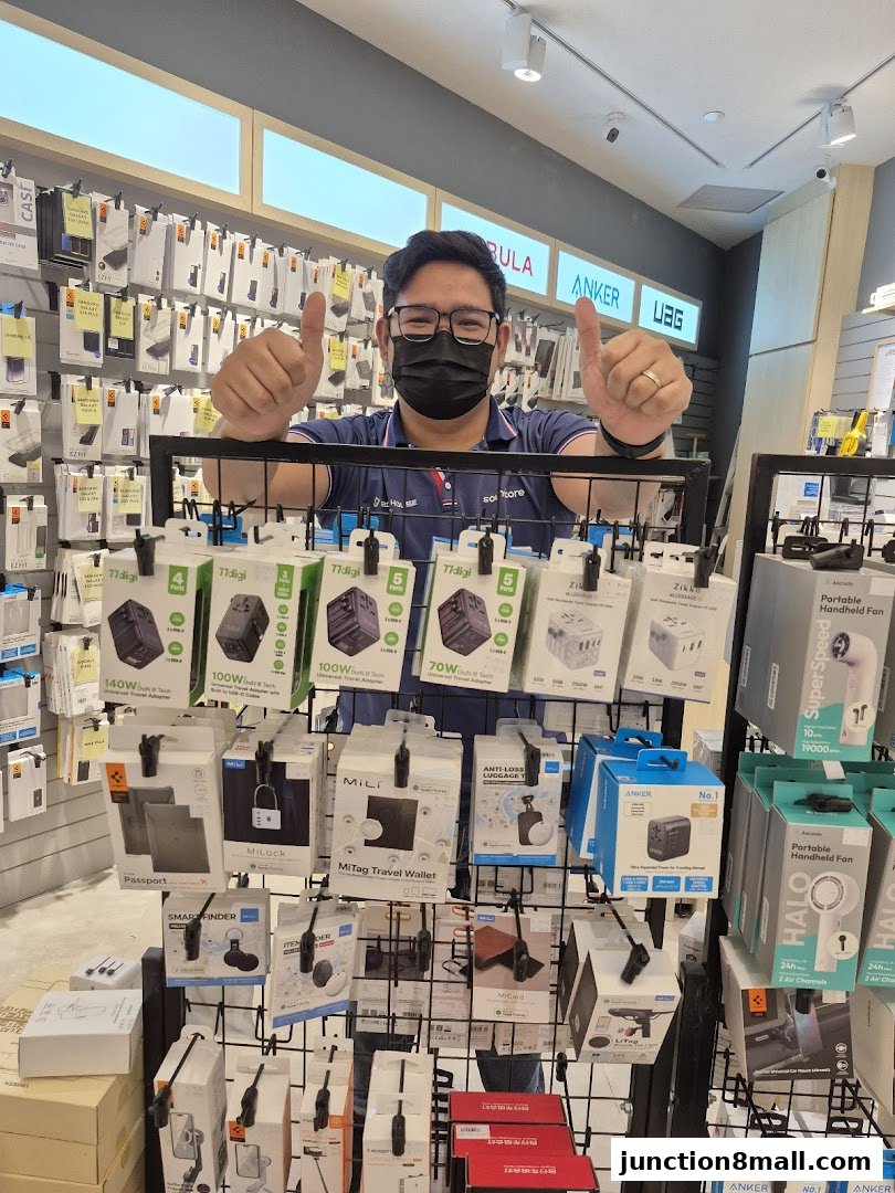 A man in a mask gives a thumbs up in front of a display of travel adapters and accessories.