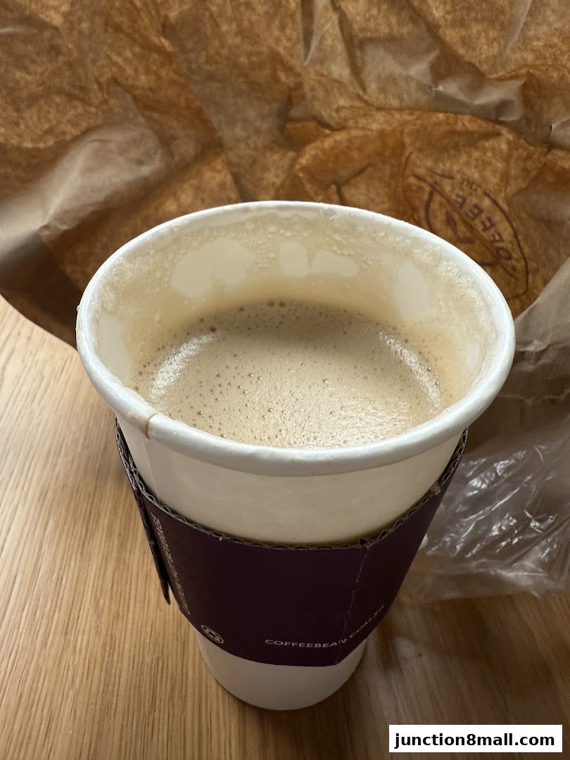 A cup of coffee with foam sits on a wooden table next to a brown paper bag.