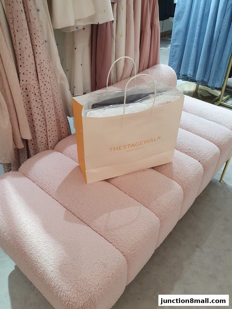 A shopping bag from The Stage Walk rests on a plush pink bench with clothing in the background.