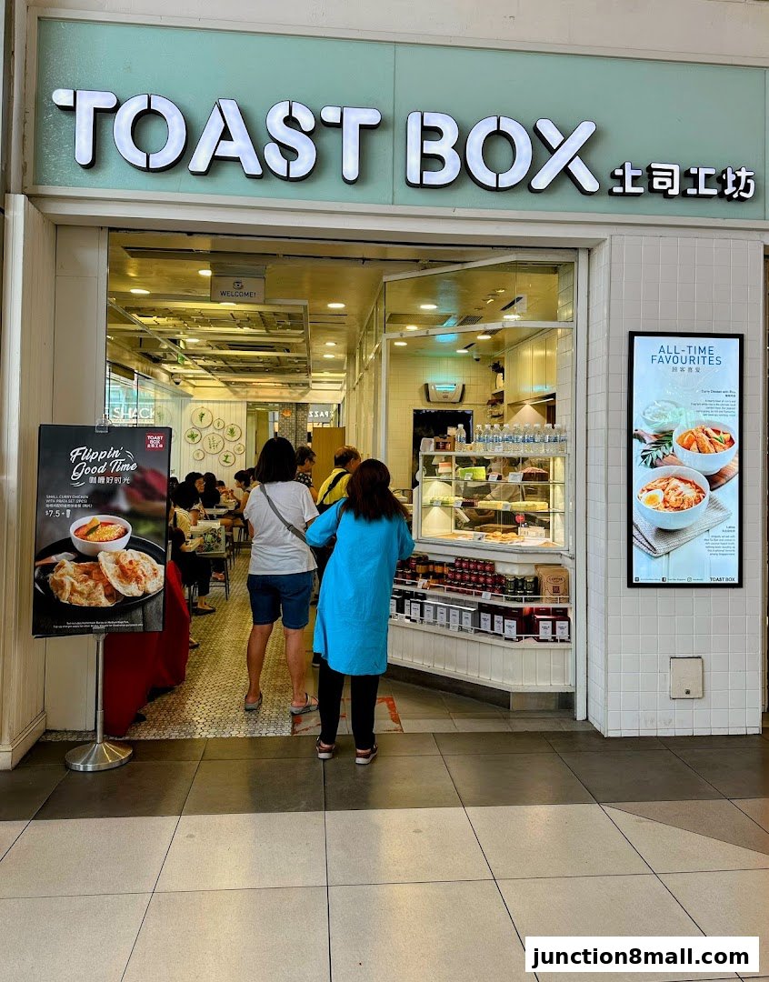 The entrance of Toast Box, a food establishment, with customers queuing and a promotional poster.