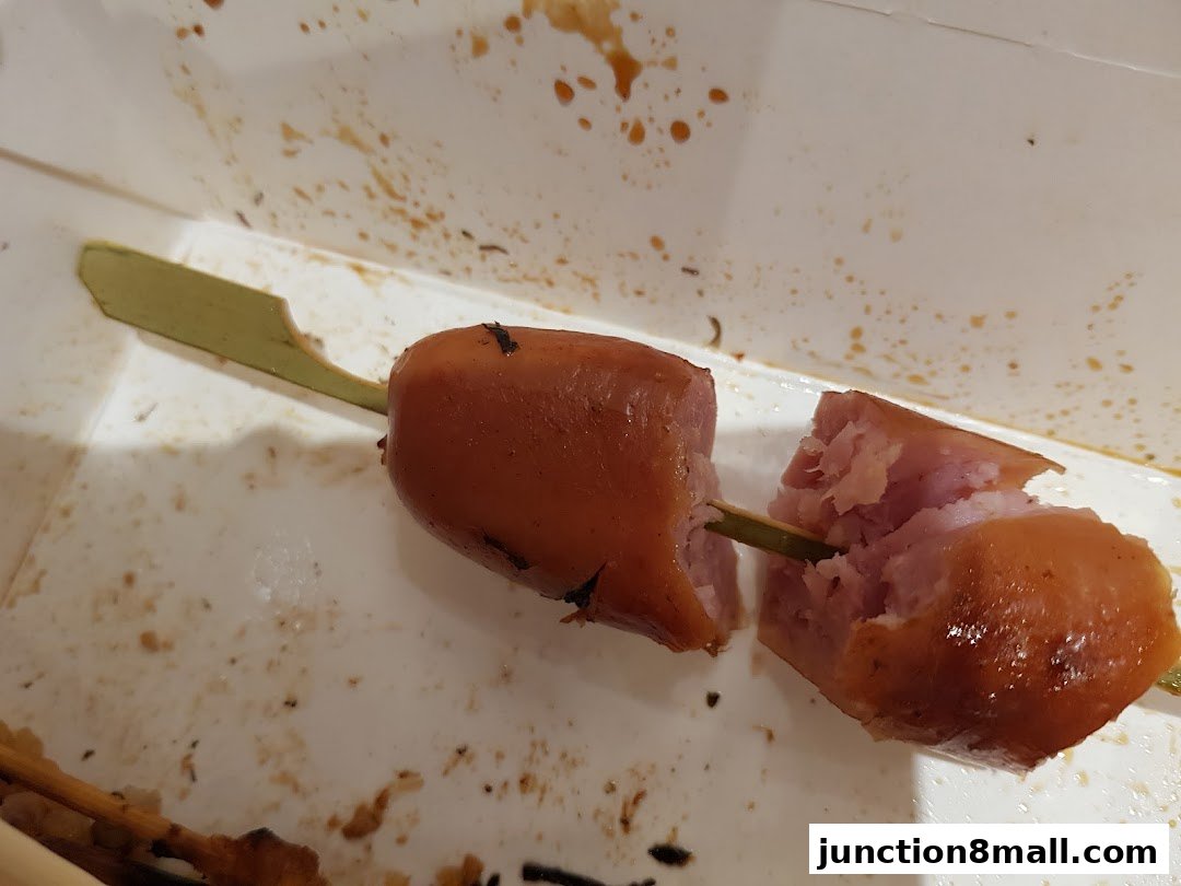 A close-up of a grilled sausage on a skewer, broken in half, in a disposable container.
