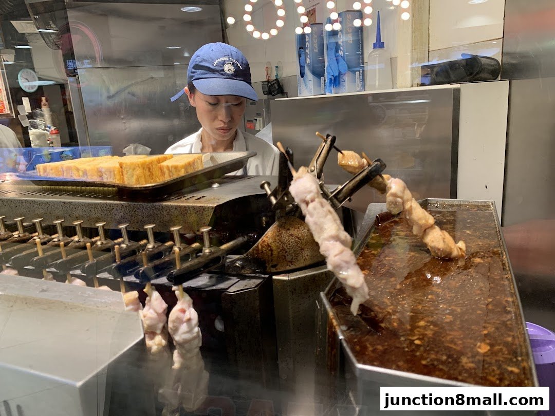 A person prepares skewers of meat over a grill with a tray of food behind them.