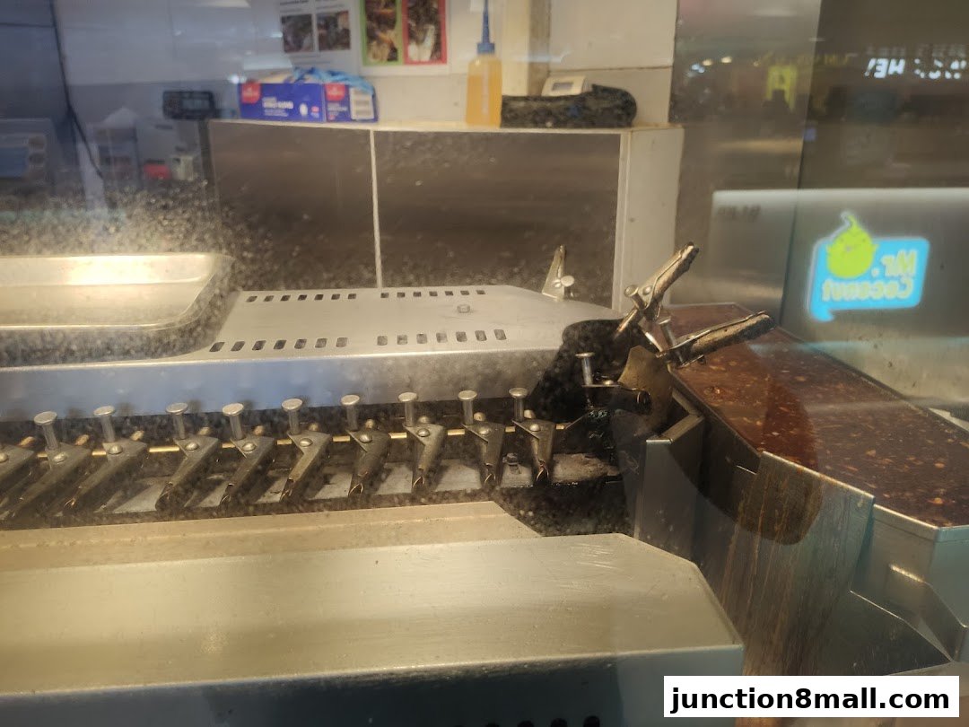 Close-up of a food preparation machine with skewers and a logo in the background.