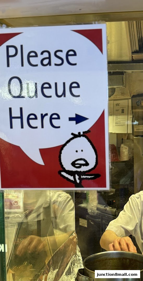 A sign indicating 'Please Queue Here' with a cartoon character and an arrow.