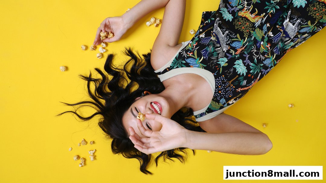 A woman in a tropical print dress lies on a yellow background, holding popcorn.