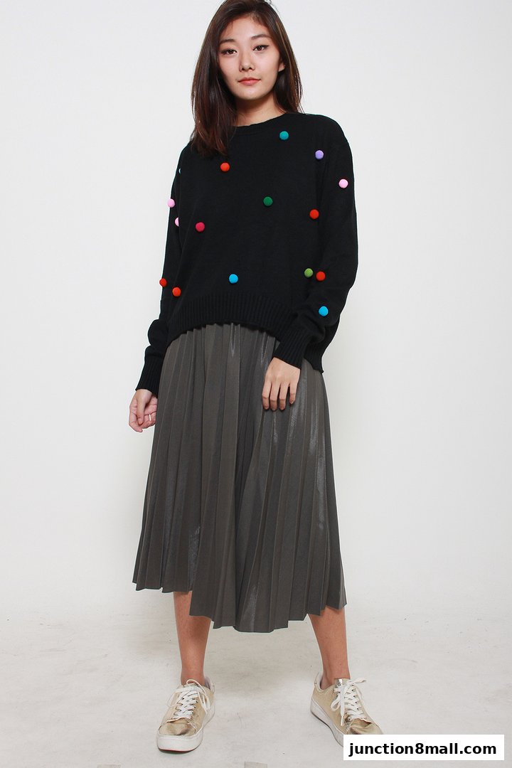 A young woman models a black sweater with colorful pom-poms and a grey pleated skirt.